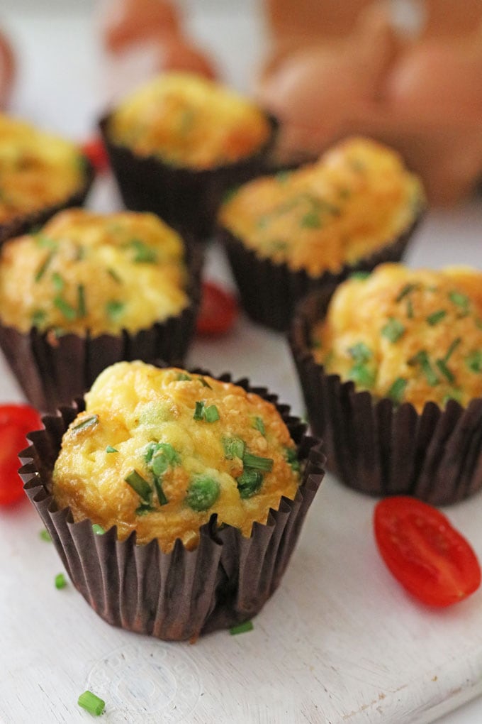 Pea & Sweetcorn Egg Muffins in brown paper muffin cases on a white chopping board with sliced cherry tomatoes.
