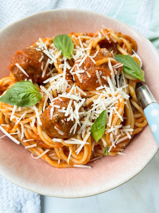 Slow cooker spaghetti meatballs served in a light pink pasta bowl, garnished with grated cheese and basil leaves. 