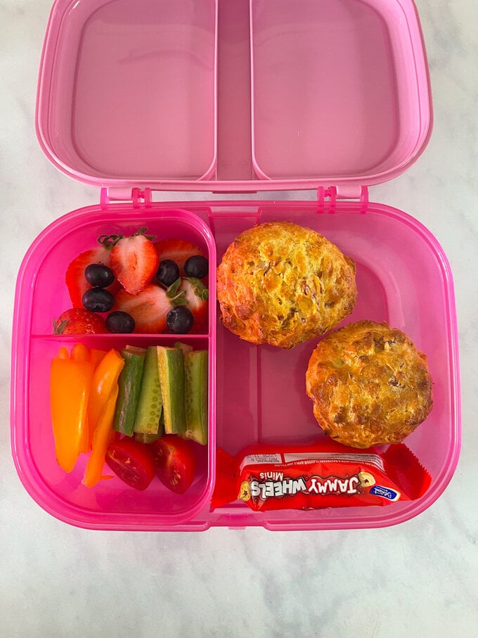 Two ham & cheese muffins served in a bright pink child's divided plastic lunchbox with fresh,chopped fruit and veggies.