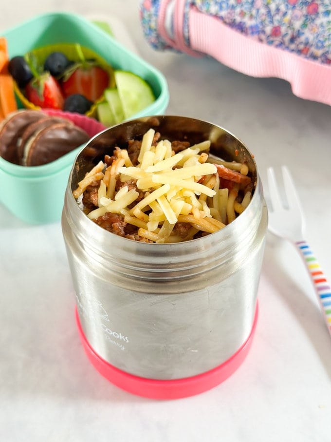 spaghetti bolognese in a children's food flask with a small green snack box in the background containing fresh fruit and veggies and some chocolate covered rice cakes.