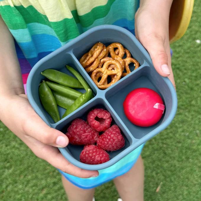 A child holding a blue silicone snack box with four compartments filled with raspberries, a cheese portion, pretzels and sugar snap peas.